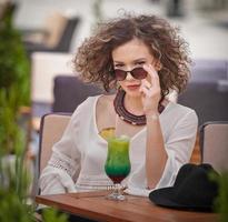 gelukkig brunette meisje met zonnebril zittend Bij de park, drinken een glas van verkoudheid groen sap terwijl glimlachen in de afstand. jong mooi vrouw Aan de bank drinken sap vervelend een wit overhemd foto