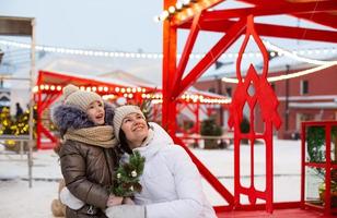 mam en dochter voor Kerstmis buitenshuis in warm kleren in winter Bij feestelijk markt. fee lichten slingers versierd sneeuw stad- voor nieuw jaar foto