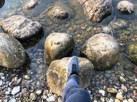 een voet in een sneaker stappen Aan groot ronde mooi natuurlijk stenen kasseien in water, zee, meer, rivier. concept toerisme, buitenshuis activiteiten foto