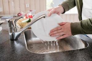 mannetje handen wassen een wit bord van voedsel overblijfselen, onder rennen water, in de wasbak, in de keuken. foto