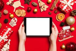 top visie van vrouw Holding tablet in haar handen Aan rood achtergrond gemaakt van Kerstmis decoraties. nieuw jaar vakantie concept. mockup foto