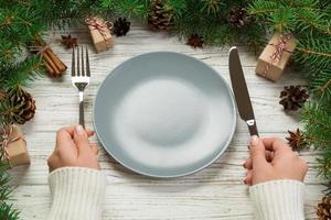 perspectief visie meisje houdt vork en mes in hand- en is klaar naar eten. leeg bord ronde keramisch Aan houten Kerstmis achtergrond. vakantie avondeten schotel concept met nieuw jaar decor foto