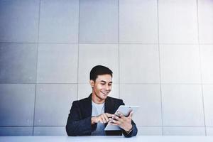 gelukkig jong Aziatisch zakenman werken Aan digitaal tablet. glimlachen en zittend Bij de bureau in industrieel zolder werkplek. breed schot foto