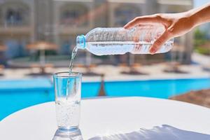 vrouw hand- gieten water in een glas van fles tegen zwemmen zwembad achtergrond foto