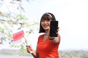 mooi jong Aziatisch vrouw draag- de Indonesisch vlag met een vrolijk gezicht en roeping zijn vriend foto
