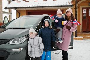 jonge noorse moeder met kinderen houdt noorse vlaggen vast en laadt elektrische auto op in de tuin van haar huis in de winter. foto