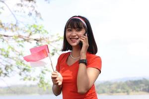 mooi jong Aziatisch vrouw draag- de Indonesisch vlag met een vrolijk gezicht en roeping zijn vriend foto