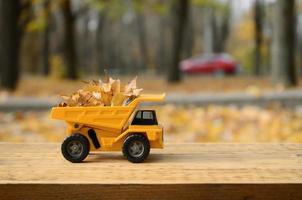 de concept van seizoensgebonden oogsten van herfst gedaald bladeren is afgebeeld in de het formulier van een speelgoed- geel vrachtauto geladen met bladeren tegen de achtergrond van de herfst park foto