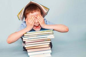 schooljongen zittend met stapel van school- boeken en covers zijn gezicht met handen geïsoleerd Aan een blauw achtergrond foto