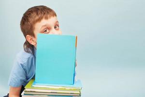 schooljongen is zittend met een stack van boeken en lezing en covers zijn gezicht met een boek geïsoleerd Aan blauw achtergrond foto