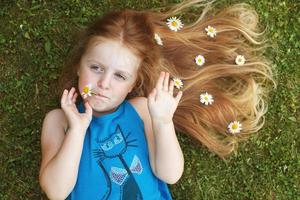 portret van een mooi weinig meisje met gezond rood haar- met kamille bloemen aan het liegen Aan de gras foto