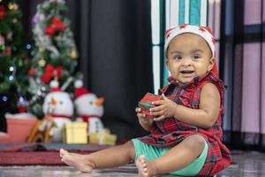 afro-amerikaanse baby lacht vrolijk terwijl hij een kleine geschenkdoos van ouders ontvangt terwijl hij zich kleedt in kerstjurk en kerstmuts met kerstboom op de rug voor het concept van de seizoensviering foto