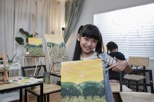 een Aziatisch leerling meisje shows haar schilderij werk, trots met portefeuille, acryl kleur afbeelding Aan canvas in kunst klas en creatief aan het leren met talenten vaardigheid Bij elementair school- studio onderwijs. foto