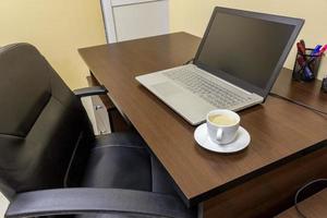 comfortabel werken plaats in kantoor met houten tafel, zwart leer stoel, laptop en kop van koffie. foto