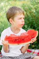 jongen aan het eten watermeloen wit t-shirt. picknick met watermeloenen. de jongen is Holding een groot stuk van watermeloen in zijn handen. helder rood sappig watermeloen. foto