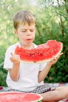 jongen aan het eten watermeloen wit t-shirt. picknick met watermeloenen. de jongen is Holding een groot stuk van watermeloen in zijn handen. helder rood sappig watermeloen. foto