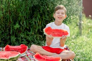 jongen aan het eten watermeloen wit t-shirt. picknick met watermeloenen. de jongen is Holding een groot stuk van watermeloen in zijn handen. helder rood sappig watermeloen. foto