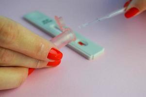 coronavirus test Aan een roze matte achtergrond. medisch procedure. een meisje met helder rood nagels voegt toe een chemisch oplossing naar de gevoelig strip naar bepalen antilichamen naar covid foto