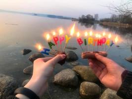 brandend gelukkig verjaardag opschrift gemaakt van vakantie kaarsen in de handen van een Mens en een vrouw tegenover de water van de oceaan meer rivier. concept verjaardag viering in natuur, buitenshuis foto