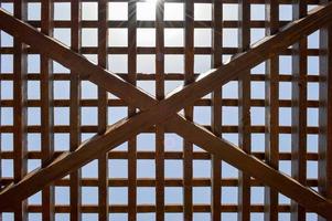 structuur van een bruin houten abstract traliewerk met plein cellen met gaten van borden van log balken geregeld verticaal horizontaal diagonaal tegen de achtergrond van de zon en lucht. de achtergrond foto