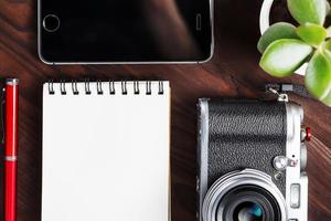 klassiek camera met blanco kladblok bladzijde en rood pen Aan donker bruin houten tafel, wijnoogst tafel met telefoon en groen bloem foto