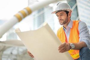 jong Kaukasisch Mens Holding een groot papier, vent vervelend licht blauw overhemd en jeans met oranje hesje en wit helm voor veiligheid in bouw Oppervlakte. foto