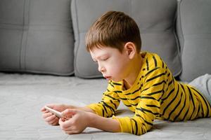 een tiener- jongen leugens Aan de bankstel met een telefoon in zijn handen. de kind Toneelstukken Bij huis in de telefoon. recreatie met de telefoon. vakantie met gadgets. foto