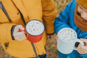 handen van jongens in een jasje houden heet cacao met marshmallows in mokken, top visie. een knus foto met een mok in hand, Aan de straat. Kerstmis humeur. kinderen drinken heet chocola Aan de straat