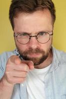 Mens met een baard en bril strikt looks en points vingers, Aan een geel achtergrond. foto