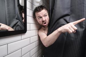 Mens is woest, gluren uit van achter een douche gordijn, hij gevangen iemand piepen Bij hem, points een vinger Bij hem en schreeuwt boos. foto