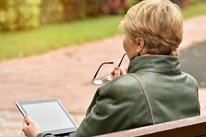 een ouderen vrouw met een tablet computer is zittend Aan een bank in een herfst park. foto