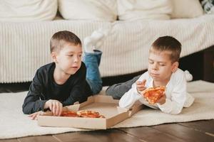 twee jongens liggen Aan de verdieping aan het eten peperoni pizza van een doos. verticaal kader. twee jongens liggen Aan de verdieping aan het eten peperoni pizza van een doos. foto
