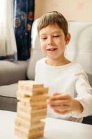 een glimlachen Kaukasisch jongen Toneelstukken Bij huis, Aan de tafel in jenga. bord spellen voor kinderen en volwassenen. een tijdverdrijf zonder gadgets. spellen gedurende de vakantie Bij huis. gelukkig kind Toneelstukken met houten blokken. foto