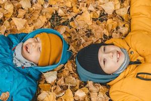 kinderen liggen Aan geel bladeren Aan de grond. jongens in jasje, verstrooit bladeren in een herfst park. de kind verheugt zich in de herfst bladeren. de jongens zijn gelukkig over herfst. gelukkig kinderjaren foto