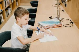 de jongen zit in de bibliotheek en schrijft in een notitieboekje Bij de tafel. voorbereidingen treffen voor huiswerk. een mooi zo leerling. schooljongen liefdes naar studie. vrij ruimte Bij school. foto