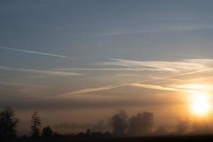 de zon stijgt achter een mist bank. de lucht gloeit blauw en oranje. de bomen verschijnen flauw in de mist. foto