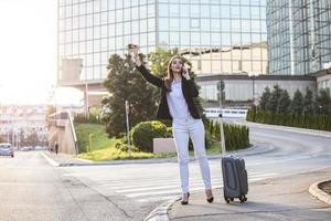 glimlachen jong vrouw probeert naar hou op taxi, roeping een taxi. jong professioneel vrouw met koffer aankoppelen een taxi in de financieel wijk, lachend. foto