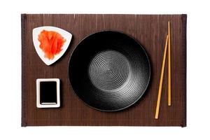 leeg ronde zwart bord met eetstokjes voor sushi, gember en soja saus Aan donker bamboe mat achtergrond. top visie met kopiëren ruimte voor u ontwerp foto