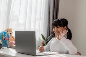 Aziatisch schoolmeisje aan het doen haar huiswerk met laptop Bij huis. kinderen gebruik gadgets naar studie. onderwijs en afstand aan het leren voor kinderen. thuisonderwijs gedurende quarantaine. blijven Bij huis foto