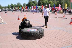 een brandweerman in een vuurvast pak en een helm loopt en bochten een groot rubber wiel in een brand vechten wedstrijd, Wit-Rusland, Minsk, 08.08.2018 foto