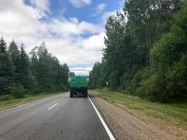 een vrachtwagen, een trekker met een groot groen aanhangwagen is het rijden langs een Woud asfalt weg met groen bomen Aan de gronden foto