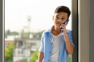 een jong jongen, een Aziatisch elementair school- leerling, is Holding een smartphone roeping en pratend naar iemand. foto
