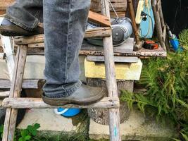 een Mens staat Aan een houten ladder in zwart rubber overschoenen. werk Bij hoogte. vuil schoenen, landbouw. tegen de achtergrond van een houten bank, plakband recorder, varen bladeren foto