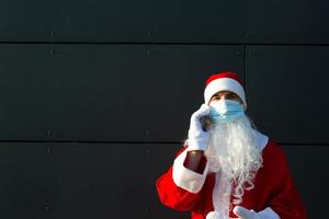 de kerstman claus in een medisch masker met een smartphone. online hartelijk groeten, bestellen Diensten voor Kerstmis en nieuw jaar. sociaal afstand in de coronavirus epidemie, een nieuw realiteit. kopiëren ruimte foto