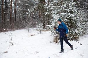 skiër met een rugzak en muts met pompon met skistokken in zijn handen op de achtergrond van een besneeuwd bos. langlaufen in het winterbos, buitensporten, gezonde levensstijl, wintersporttoerisme. foto