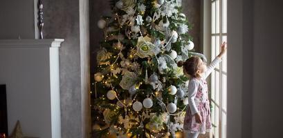 een weinig meisje looks uit van een groot venster in de buurt een Kerstmis boom. aan het wachten voor een wonder, Kerstmis wit decor in de leven kamer van de huis. nieuw jaar, fee verhaal en magie, kinderen dromen foto