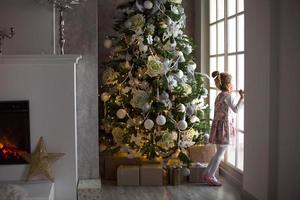 een weinig meisje looks uit van een groot venster in de buurt een Kerstmis boom. aan het wachten voor een wonder, Kerstmis wit decor in de leven kamer van de huis. nieuw jaar, fee verhaal en magie, kinderen dromen foto