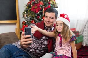 vader en dochter in de buurt de Kerstmis boom met een smartphone nemen een selfie, communiceren via video verbinding. Kerstmis hartelijk groeten, een geschenk doos, een meisje in een de kerstman hoed golvend Hallo. nieuw jaar, vakantie decor foto