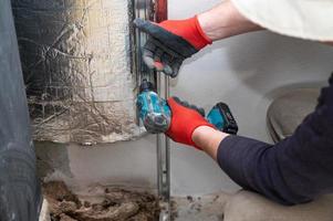 de meester schroeven een metaal profiel naar de muur met een schroevendraaier, opwarming de muur in de buurt de haard met mineraal wol en folie. foto