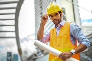 jong Kaukasisch Mens Holding een groot papier, vent vervelend licht blauw overhemd en jeans met oranje hesje en geel helm voor veiligheid in bouw Oppervlakte. foto